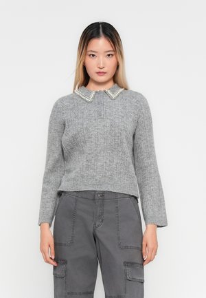 Young woman with straight ombre hair wearing a grey knit sweater with pearl-trimmed collar and grey cargo pants standing against white background.
