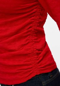 Rød teksturert stofftopp med samlet sidesøm, iført mørkeblå denimjeans, som viser midje- og midtseksjonsdetaljer.