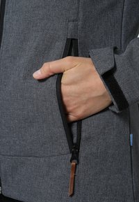 Veste grise en tissu texturé, équipée de poches zippées et d'une languette en cuir. Les poignets sont dotés d'un accent Velcro pour un ajustement réglable.