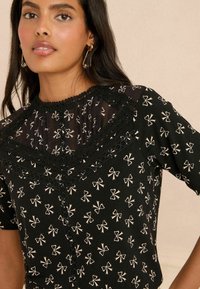 Blouse noire avec un empiècement transparent plissé, en tissu à motif nœud, garniture en dentelle et boutons décoratifs sur le devant.
