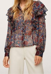 Femme portant un chemisier transparent à manches longues avec des épaules volantes et un motif paisley floral multicolore, associé à un pantalon beige clair.