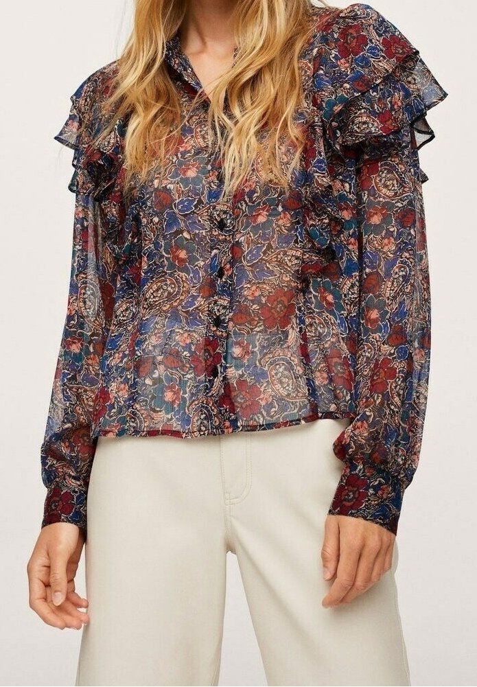 Femme portant un chemisier transparent à manches longues avec des épaules volantes et un motif paisley floral multicolore, associé à un pantalon beige clair.