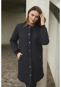 Manteau noir matelassé avec une fermeture à boutons, poches latérales et poignets côtelés. Présente un motif en losange sur le tissu et une coupe mi-longue.