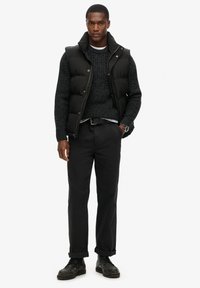 Chaleco acolchado negro sobre un suéter de punto oscuro, combinado con pantalones negros remangados en los puños y zapatos de cuero negros. Diseño minimalista.