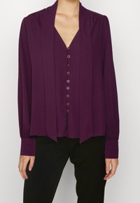 Kobieta w koszuli z długim rękawem w kolorze śliwkowym, z złotymi guzikami z przodu, w połączeniu z czarnymi spodniami na jednolitym tle.