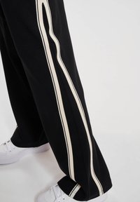 Svarte bukser med brede hvite vertikale striper, iført med hvite sneakers på lys bakgrunn.