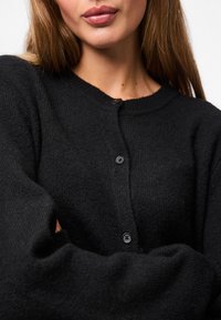 Cardigan en tricot noir avec un col rond, doté d'une fermeture à boutons et de manches longues. Texture douce et coupe décontractée.