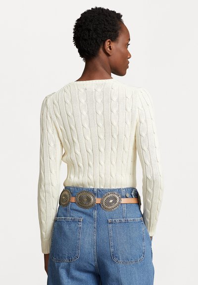Person med kort krøllet hår iført en hvid kabelstrikket sweater og højtaljede blå jeans med et dekorativt bælte.