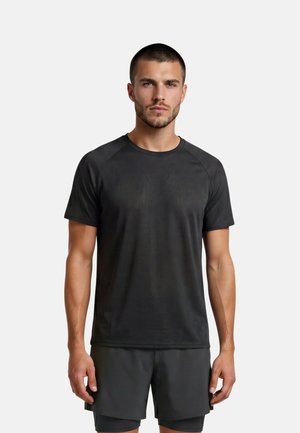 Man met kort haar die een eenvoudige zwarte sport-T-shirt met korte mouwen en bijpassende zwarte korte broek draagt, staand tegen een witte achtergrond.