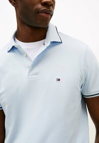 Camisola polo azul clara feita de tecido texturizado, com um colarinho em contraste e um pequeno logótipo em vermelho, branco e azul no peito.