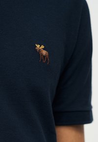 Ναυτικό μπλε ύφασμα με μικρό κεντημένο καφέ λογότυπο ελαφιού με κίτρινα κέρατα στο πάνω μέρος του στήθους ενός ενδύματος με κοντά μανίκια.