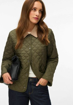 Kvinde iført olivengrøn quilted jakke med beige krave, mørke jeans, holder en sort clutch taske, ser til siden.