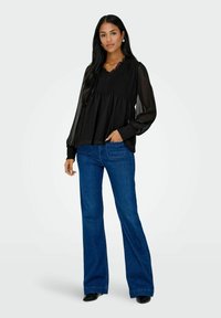 Femme portant un chemisier noir transparent à manches longues et un jean évasé bleu, debout avec une main dans sa poche et des chaussures noires.