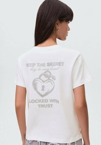 T-shirt en coton blanc avec un design graphique au dos : un cœur avec un trou de serrure et du texte. Manches courtes, coupe décontractée, texture lisse.