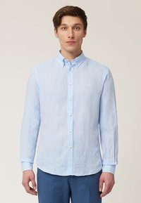 Chemise bleu clair à manches longues avec boutons, en tissu texturé, présentant un col classique, des boutons sur le devant et un petit logo sur la poitrine.