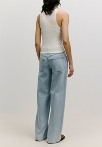 Weißes geripptes Tanktop kombiniert mit hellblauen weit geschnittenen Jeans mit zwei Gesäßtaschen; das Model steht mit dem Rücken zur Kamera.
