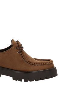 Zapato de ante marrón con un diseño estilo mocasín, punta redonda, costuras contrastantes y una suela gruesa y robusta de goma negra. Se pueden ver dos ojales.