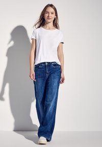 T-shirt bianco a maniche corte abbinato a jeans a gamba larga blu, con un classico design a cinque tasche e chiusura con bottone e cerniera.