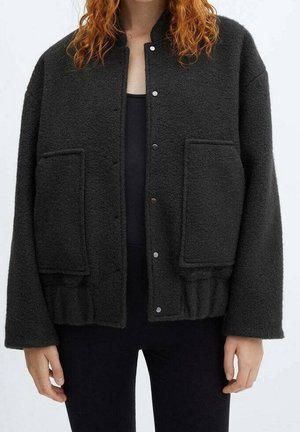 Personne portant une veste noire à boutons-pression avec de grandes poches avant, par-dessus un haut noir et un pantalon noir, montrée du cou jusqu’aux hanches.