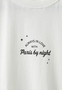 Bela bombažna majica z natisnjenim črnim napisom: "VEDNO ZALJUBLJEN V Pariz ponoči," poudarjen z majhnimi zvezdicami okoli besedila.