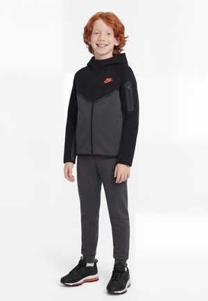 Rudy chłopiec uśmiechający się, ubrany w czarną i szarą bluzę z kapturem Nike, dopasowane dresy i czarne trampki z czerwonymi i białymi podeszwami.