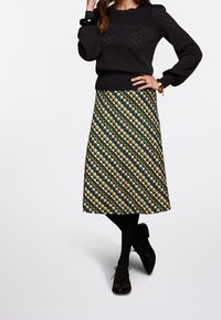 Musta neulepusero, jossa on teksturoitu sydänkuvio, yhdistetty vihreään ja keltaiseen kuvioituun puolihameeseen, jossa on vinoja raitoja. Mustat leggingsit ja kengät.