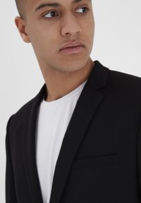 Blazer negro de tela suave, con solapas en pico, un bolsillo y usado sobre una camiseta blanca de cuello redondo.