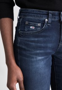 Jeans de mezclilla azul oscuro usados por una persona con piel de tono medio oscuro, mostrando el bolsillo delantero con un pequeño parche rectangular de logo.
