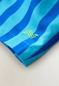 Nahaufnahme eines blau gestreiften Stoffs mit einem genähten Saum und einem kleinen gelben, gestickten Wellensymbol.