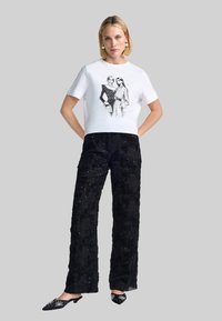 Weißes, kurz geschnittenes T-Shirt mit Frontillustration, kombiniert mit schwarzen, mit Pailletten besetzten, weit geschnittenen Hosen. Schwarze, veredelte Mules runden das Outfit ab.