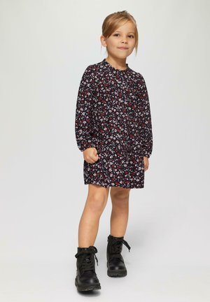 Sukienka w kwiaty w kolorze granatowym z długimi rękawami i falbaniastym dekoltem, zestawiona z czarnymi butami na sznurówki. Sylwetka w kształcie litery A.