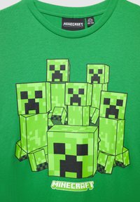 T-shirt verde in cotone con design pixelati di creeper di Minecraft in diverse sfumature di verde, con il logo "Minecraft" stampato in bianco nella parte inferiore.