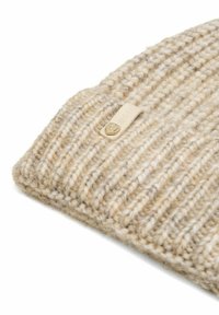 Szydełkowana czapka w jasnym beżu z wyraźną strukturą ribbed. Posiada małą, złotą metalową metkę. Materiał wydaje się miękki i gruby.