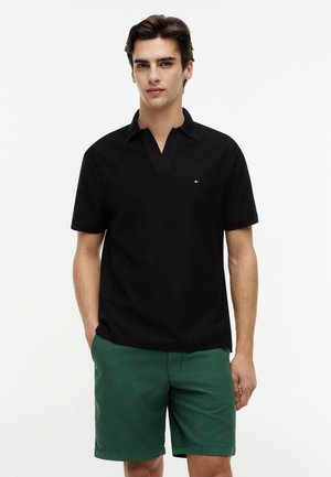 Młody mężczyzna stojący, ubrany w czarną koszulkę polo z krótkim rękawem oraz zielone spodenki, z rękami częściowo wsuniętymi do kieszeni, na białym tle.