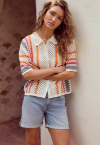 Gilet en tricot avec un design ajouré en blanc, orné de rayures orange, rose et grise. Associé à un short en denim roulé bleu clair.