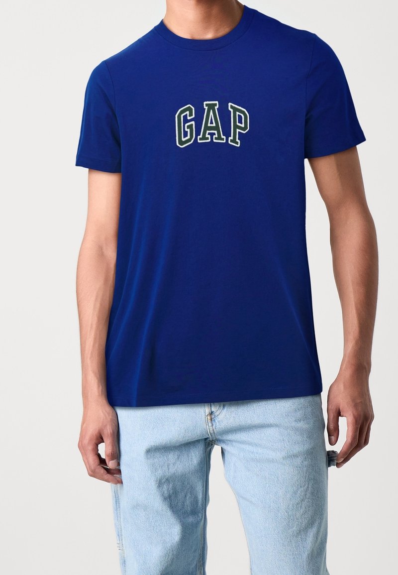 Blue cotton t-shirt with short sleeves, featuring a white and green "GAP" logo on the front. Paired with light blue denim jeans.