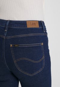 Mörkblå denimjeans med en tan läderlogotyp, kontrasterande gula sömmar och design av rundad bakficka.