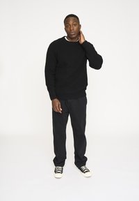 Sort tekstureret striksweter, sorte løstsiddende bukser og sort-hvide sneakers. Model står op ad en simpel hvid baggrund.