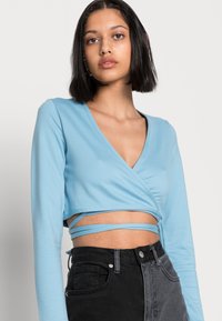 Top cropped de manga longa azul claro com um design envolvente, apresentando tiras finas à volta da cintura, combinado com jeans pretos de cintura alta.