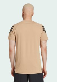 Tan athletic t-shirt with a textured surface, short sleeves, and black accent stripes on the shoulders. Features side slits at the hem.
