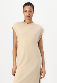 Robe midi beige à manches courtes, fabriquée dans un tissu doux. Elle présente un col rond et un logo discret sur la poitrine. Texture lisse.