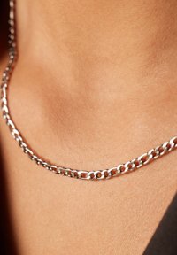 Gros plan d'un collier en argent reposant sur une peau lisse à la base du cou, avec un aperçu de vêtements noirs visibles.