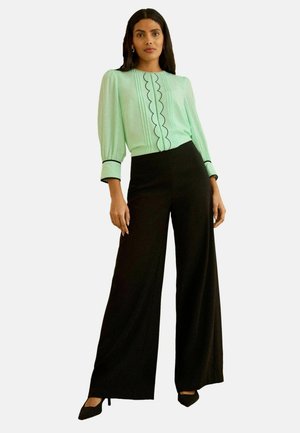 Femeie purtând o bluză verde mentă cu margine neagră ondulată și pantaloni negri largi, stând în picioare pe un fundal simplu.