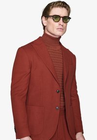 Rød skræddersyet blazer med sorte knapper, båret over en struktureret rød turtleneck. Tilbehøret inkluderer briller med grønlig tone. Simpel baggrund.