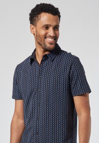 Lyhythelmainen paita tummansinisessä värissä, jossa on vaaleansiniset geometriset kuviot. Napillinen etuosa, kaulus ja rento leikkuu. Sujuva kankaan pinta.