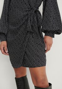 Robe portefeuille noire à pois blancs avec manches longues bouffantes, associée à des bottes noires texturées montant jusqu'au genou.