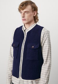 Jūras spēku zīmola polāra veste ar krējuma krāsas rāvējslēdzēju un apdari. Aprīkota ar divām priekšējām kabatām ar brūniem pogām. Valkāta virs šūpuļa krekla ar pārbaudītu rakstu.