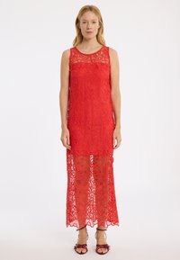 Rotes Spitzenkleid mit Blumenmustern, ärmellos und mit einem durchsichtigen Saum. Kombiniert mit passenden roten Riemchensandalen.