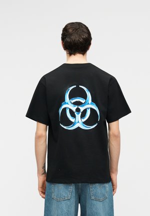 Άνδρας που φοράει μαύρο μπλουζάκι με μεγάλο μπλε σύμβολο βιολογικού κινδύνου στην πλάτη, σε συνδυασμό με ανοιχτό μπλε τζιν, στέκεται μπροστά σε απλό φόντο.