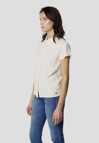Blusa de manga corta color crema claro con cierre de botones en la parte delantera, tela texturizada, detalles de bordado floral y diseño de cuello clásico.